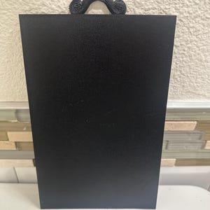 May include: A black rectangular sign with a textured surface and a decorative handle at the top. The handle has a swirled design. The sign is displayed against a neutral background.
