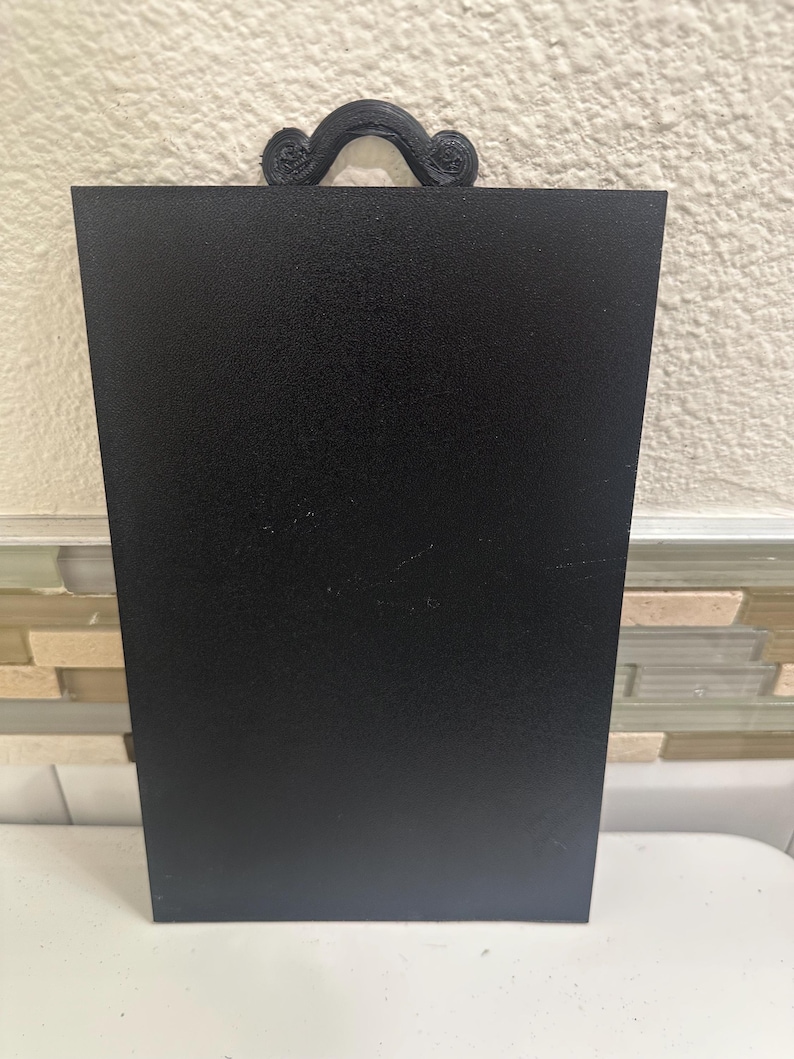 May include: A black rectangular sign with a textured surface and a decorative handle at the top. The handle has a swirled design. The sign is displayed against a neutral background.