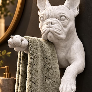 Könnte beinhalten: Ein weißer, gesprenkelter Handtuchhalter in Form einer französischen Bulldogge zur Wandmontage. Die Skulptur zeigt den Kopf und die Vorderpfoten des Hundes, wobei eine Pfote ein gefaltetes, strukturiertes salbeigrünes Handtuch hält.
