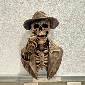 Może przedstawiać: Brązowa, montowana na ścianie rzeźba szkieletu w kapeluszu i płaszczu, trzymającego cygaro. Rzeźba ma szczegółowe żebra i rysy twarzy. Tło to teksturowana biała ściana.