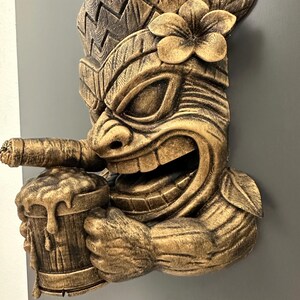 May include: A detailed, golden-brown tiki mask with a smiling face, holding a mug of beer and a cigar. The mask features a flower on its head and intricate carved details, creating a tropical, Polynesian theme.