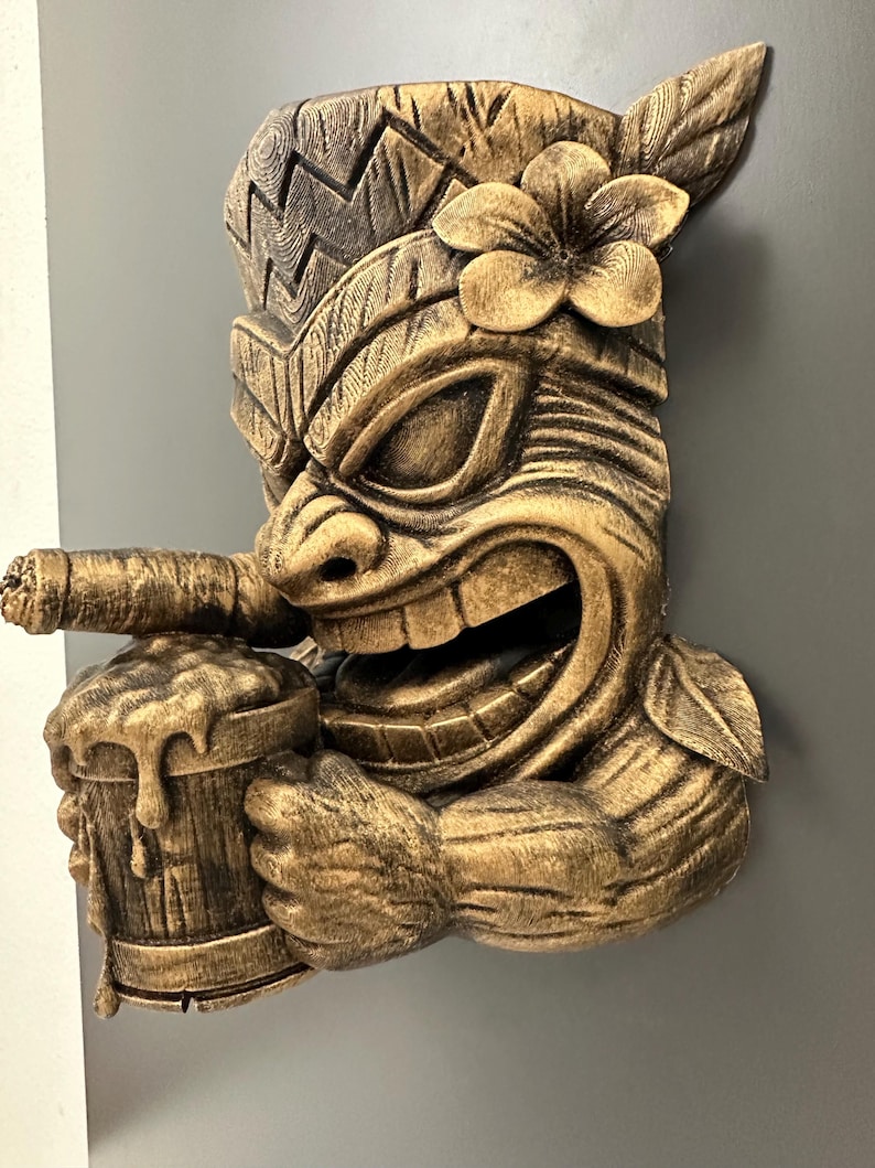 May include: A detailed, golden-brown tiki mask with a smiling face, holding a mug of beer and a cigar. The mask features a flower on its head and intricate carved details, creating a tropical, Polynesian theme.