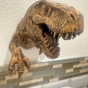 May include: A brown, textured Tyrannosaurus Rex head wall decor with an open mouth revealing sharp teeth. The dinosaur head has a detailed, realistic design, including the texture of the skin and the shape of the eye sockets. The clawed arm extends from the head.