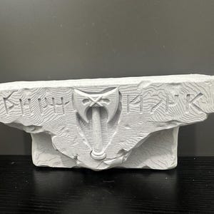 Pode incluir: Uma escultura em pedra cinza claro esculpida com uma superfície texturizada. A escultura apresenta um design de machado e runas. A pedra é retangular com uma borda áspera e natural. A escultura é exibida em uma superfície preta.