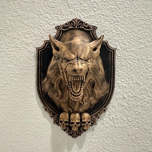 May include: A bronze-colored wall plaque featuring a snarling werewolf head with exposed fangs. The plaque has a decorative frame with three small skulls at the bottom. The background is a textured white wall.