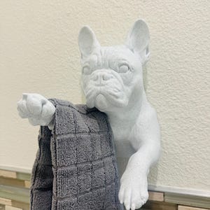 Puede incluir: Un toallero de bulldog francés blanco con una toalla gris. La escultura del perro está montada en una pared y tiene una apariencia texturizada. La toalla está doblada y tiene un patrón cuadrado.