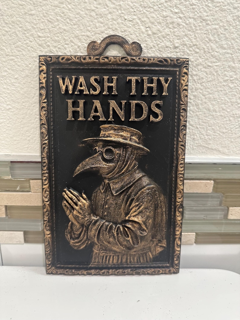 May include: A rectangular wall plaque with the words "WASH THY HANDS" in gold lettering. The plaque features a relief image of a plague doctor in a hat and mask, with a decorative frame. The background is black.