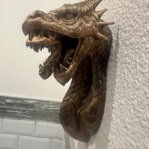 May include: A detailed, bronze-colored dragon head wall sculpture with an open mouth, sharp teeth, and textured scales. The dragon's head is mounted on a textured white wall, showcasing its intricate design and mythical theme.