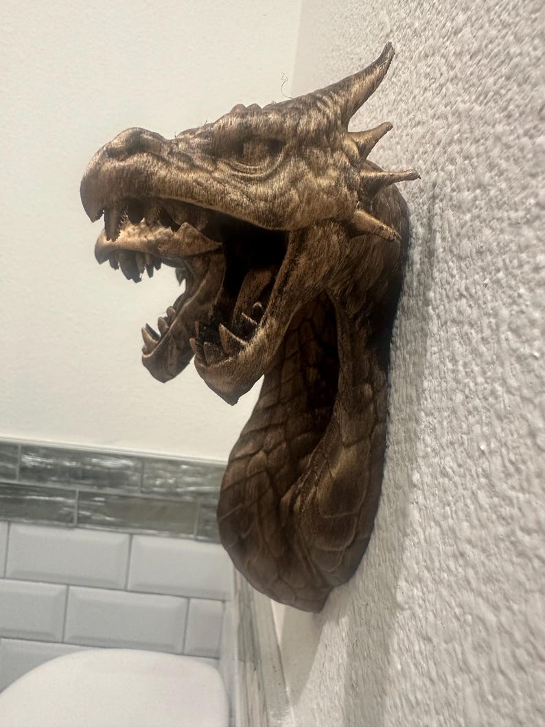 May include: A detailed, bronze-colored dragon head wall sculpture with an open mouth, sharp teeth, and textured scales. The dragon's head is mounted on a textured white wall, showcasing its intricate design and mythical theme.