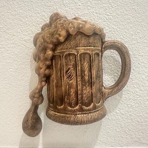 May include: A brown, textured beer stein wall hanging with overflowing foam. The stein has a handle and vertical grooves, giving it a rustic appearance. The foam drips down the side, creating a dynamic, three-dimensional effect.