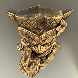 May include: A gold-colored, carved tiki wall mask with a detailed design. The mask features a stylized face with a cigar, a flower, and leaf accents. The mask is mounted on a gray wall.