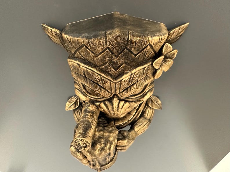 May include: A gold-colored, carved tiki wall mask with a detailed design. The mask features a stylized face with a cigar, a flower, and leaf accents. The mask is mounted on a gray wall.