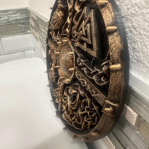 May include: A round, black and gold-colored decorative shield with intricate relief carvings. The shield features a central boss and is edged with gold-colored spikes. The carvings depict various figures and symbols, creating a detailed and textured appearance.