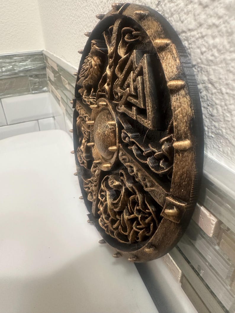 May include: A round, black and gold-colored decorative shield with intricate relief carvings. The shield features a central boss and is edged with gold-colored spikes. The carvings depict various figures and symbols, creating a detailed and textured appearance.