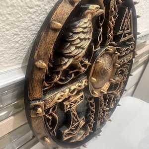 May include: A round, black and gold decorative shield with a detailed relief design. The shield features a central raven, a hammer, and other Norse symbols. The outer edge has gold-colored spikes.