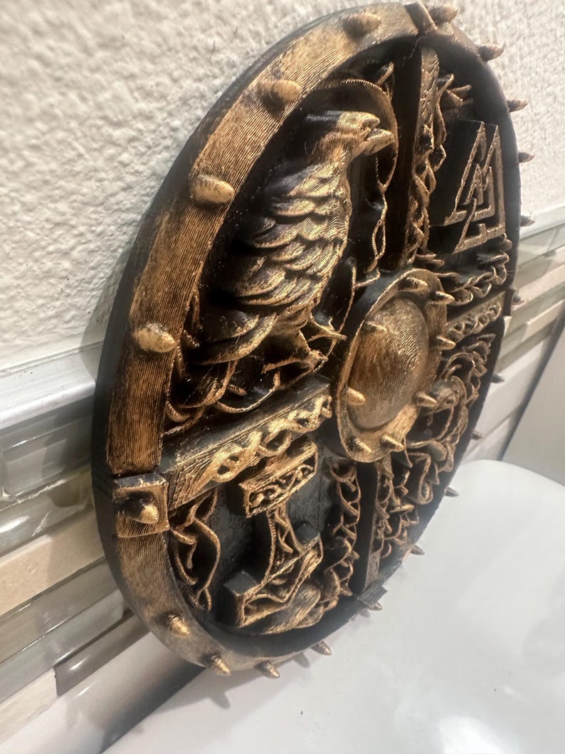 May include: A round, black and gold decorative shield with a detailed relief design. The shield features a central raven, a hammer, and other Norse symbols. The outer edge has gold-colored spikes.