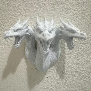 May include: A three-headed dragon wall sculpture in speckled white. Each dragon head has open jaws, sharp teeth, and detailed scales. The sculpture is mounted on a textured, off-white wall, creating a dramatic display.