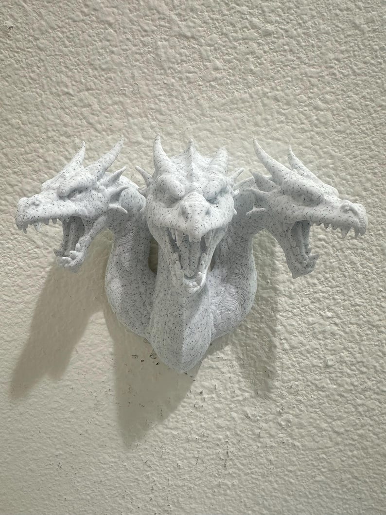 May include: A three-headed dragon wall sculpture in speckled white. Each dragon head has open jaws, sharp teeth, and detailed scales. The sculpture is mounted on a textured, off-white wall, creating a dramatic display.
