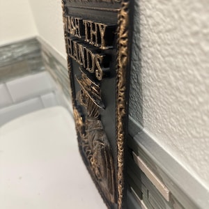 May include: A rectangular black and gold wall plaque with the words "WASH THY HANDS" in gold lettering. The plaque features a decorative border and a relief image of a figure in a long coat and hat.
