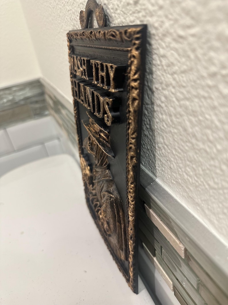 May include: A rectangular black and gold wall plaque with the words "WASH THY HANDS" in gold lettering. The plaque features a decorative border and a relief image of a figure in a long coat and hat.