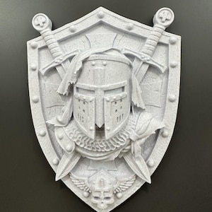 Peut inclure: Un bouclier gris clair imprimé en 3D représentant un casque de chevalier, des épées croisées et un emblème de crâne. Le bouclier a une surface texturée et est monté sur un fond sombre. Le design est détaillé et complexe.