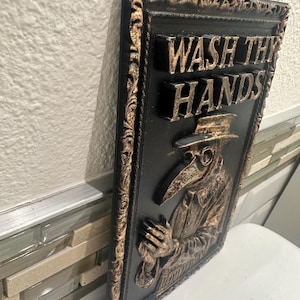 May include: Black and gold rectangular wall plaque with the words "WASH THY HANDS" in raised lettering. The plaque features a relief image of a plague doctor. The frame and lettering have a gold finish.