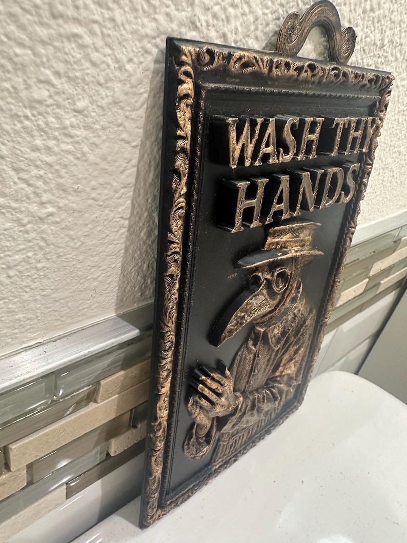 May include: Black and gold rectangular wall plaque with the words "WASH THY HANDS" in raised lettering. The plaque features a relief image of a plague doctor. The frame and lettering have a gold finish.