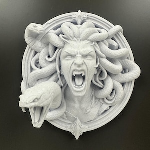 May include: A gray, 3D-printed sculpture of Medusa's head, featuring snakes and an open mouth. The sculpture is round with a decorative border and is set against a dark background.