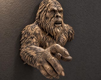May include: A wall-mounted sculpture of a brown, hairy creature resembling a Bigfoot or Sasquatch. The figure features a detailed face with an open mouth, textured fur, and an outstretched arm with a hand reaching forward in a grasping motion.