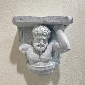 May include: A grey, textured wall shelf featuring a sculpted figure of a muscular man supporting a rectangular surface. The shelf has a classical design with intricate details, ideal for displaying small items.