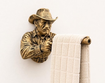 May include: A bronze-colored cowboy-themed towel holder. The holder is a sculpted bust of a cowboy wearing a hat and shirt, holding a wooden rod with a beige towel. The towel has a checkered pattern.