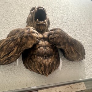 May include: A detailed, brown wall sculpture of a roaring bear with its arms raised. The bear's muscular chest and arms are prominently displayed, with intricate carved details. The sculpture is mounted on a textured, off-white wall.