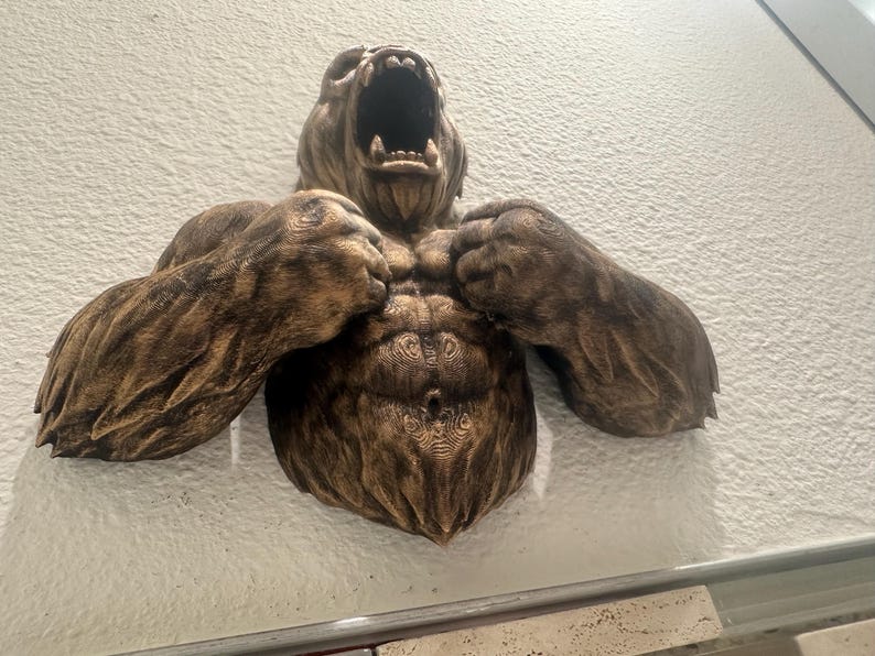 May include: A detailed, brown wall sculpture of a roaring bear with its arms raised. The bear's muscular chest and arms are prominently displayed, with intricate carved details. The sculpture is mounted on a textured, off-white wall.