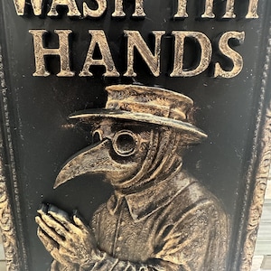 May include: A black and gold relief plaque with the text "WASH THY HANDS" above an image of a plague doctor. The doctor is wearing a hat and a long-beaked mask. The plaque has a decorative border.