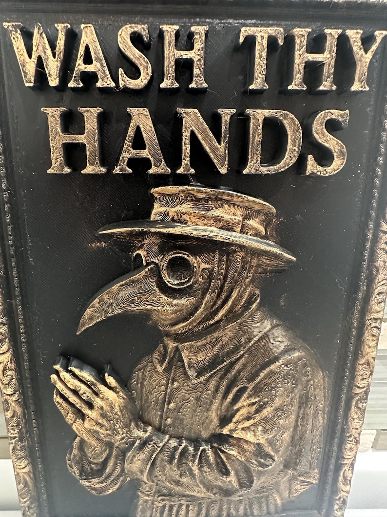May include: A black and gold relief plaque with the text "WASH THY HANDS" above an image of a plague doctor. The doctor is wearing a hat and a long-beaked mask. The plaque has a decorative border.