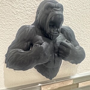 May include: A black gorilla bust sculpture with an open mouth, showcasing detailed musculature. The sculpture is mounted on a textured white wall, creating a striking contrast. The gorilla's fierce expression and powerful physique are prominently displayed.