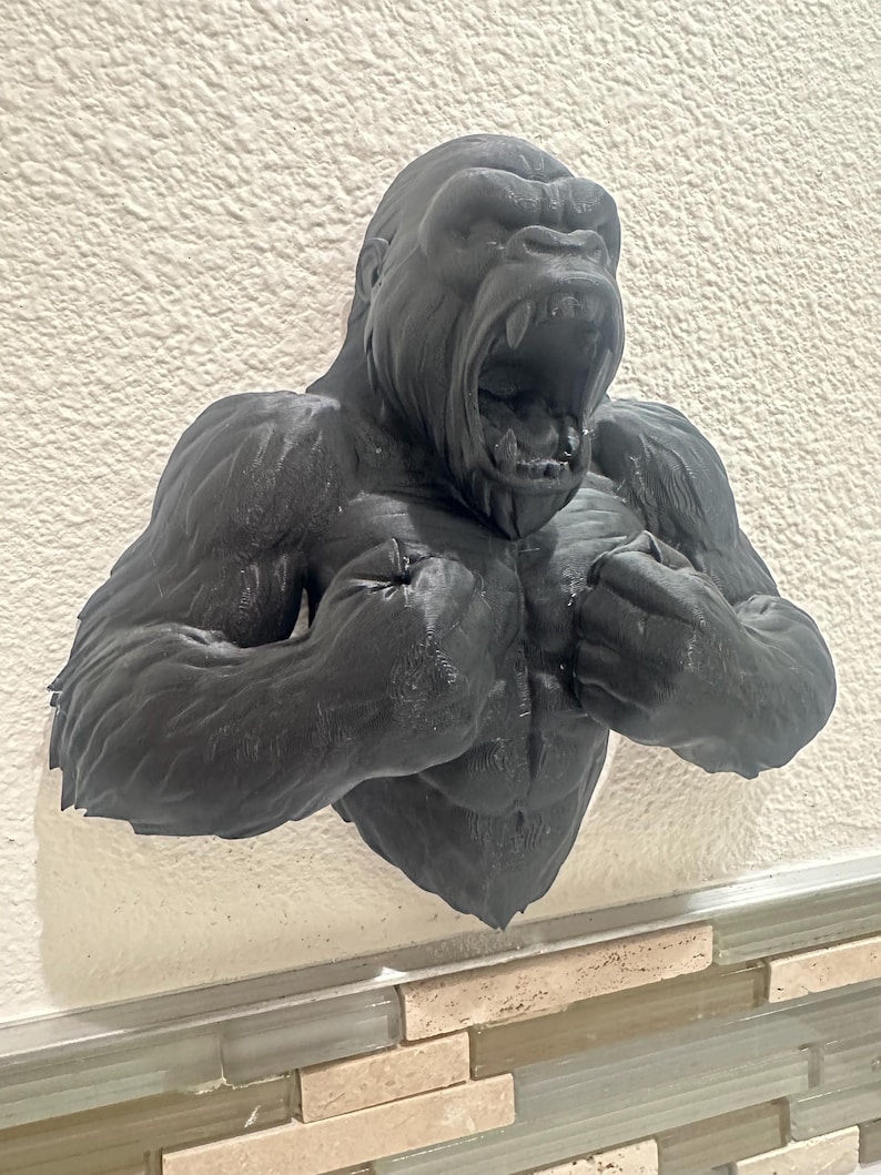 May include: A black gorilla bust sculpture with an open mouth, showcasing detailed musculature. The sculpture is mounted on a textured white wall, creating a striking contrast. The gorilla's fierce expression and powerful physique are prominently displayed.