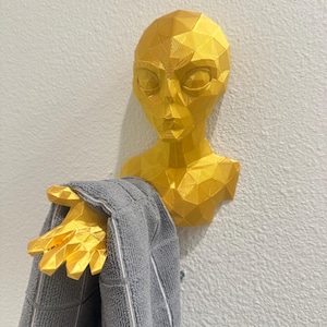 May include: A gold-colored, geometric alien head wall hook holding a gray towel. The alien head has large, almond-shaped eyes and a triangular face. The towel is gray with a white border.