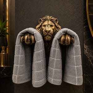 Puede incluir: Un toallero de pared con una cabeza de león esculpida en tono dorado y dos patas extendidas. Las garras del león están posicionadas para sujetar dos toallas grises texturizadas, creando un soporte funcional para ropa de baño.