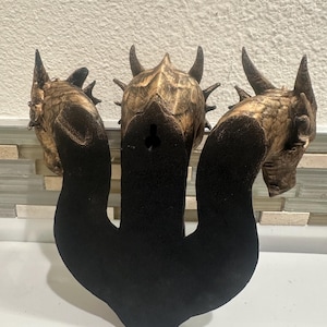 May include: A wooden wall hanging featuring three dragon heads with horns and textured details. The dragon heads are brown and gold, with a black base. The piece is designed to be mounted on a wall.