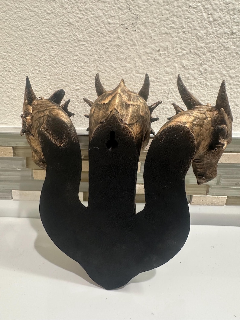 May include: A wooden wall hanging featuring three dragon heads with horns and textured details. The dragon heads are brown and gold, with a black base. The piece is designed to be mounted on a wall.