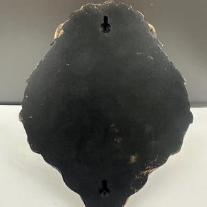 K&ouml;nnte beinhalten: Eine schwarze, schildf&ouml;rmige Wandplatte mit gewelltem Rand und goldfarbenem Finish entlang der Kante. Die R&uuml;ckseite verf&uuml;gt &uuml;ber zwei vertikal angeordnete Schl&uuml;sselloch-Aufh&auml;ngungen, die eine sichere Befestigung an der Wand erm&ouml;glichen.