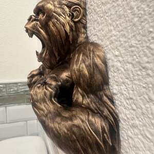 May include: A bronze-colored gorilla sculpture with an open mouth, mounted on a textured white wall. The sculpture is detailed, showing the gorilla's facial features and fur texture. The gorilla's arms are flexed, and the sculpture is partially visible.