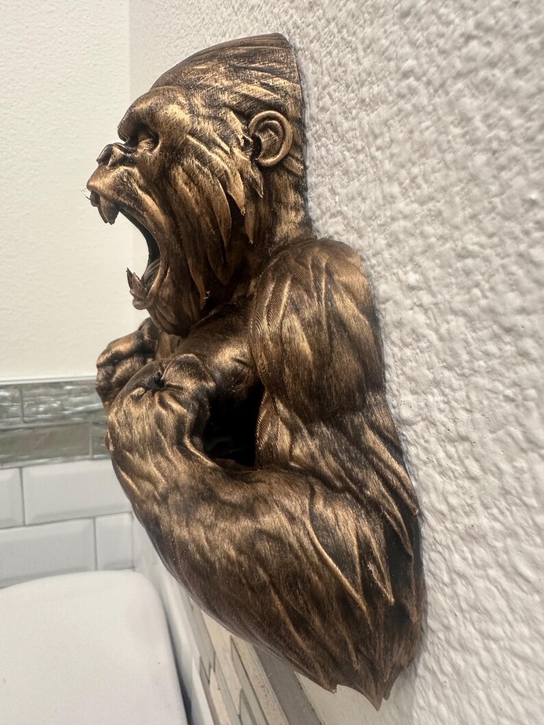 May include: A bronze-colored gorilla sculpture with an open mouth, mounted on a textured white wall. The sculpture is detailed, showing the gorilla's facial features and fur texture. The gorilla's arms are flexed, and the sculpture is partially visible.
