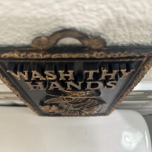 May include: A decorative black and gold sign with the words "WASH THY HANDS" in gold lettering. The sign has a handle at the top and a detailed border. The bottom of the sign features a detailed image.