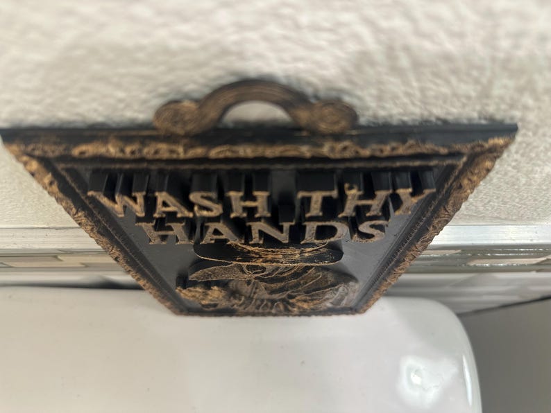May include: A decorative black and gold sign with the words "WASH THY HANDS" in gold lettering. The sign has a handle at the top and a detailed border. The bottom of the sign features a detailed image.