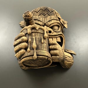 May include: A carved wooden tiki mask with a beer stein. The mask has a wide grin and is holding a wooden mug overflowing with dripping liquid. The tiki is brown with carved details and is a decorative wall hanging.