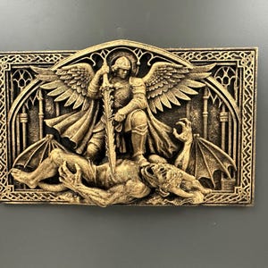 May include: A rectangular, gold-colored relief sculpture depicting a winged figure with a sword standing over a defeated demon. The sculpture has intricate details and is set against a dark background.
