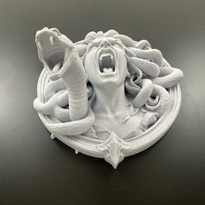 May include: A white sculpture of Medusa with snakes for hair, screaming with her mouth open. A snake is coiled around her head, with its mouth open. The sculpture is mounted on a circular base with a decorative bottom.
