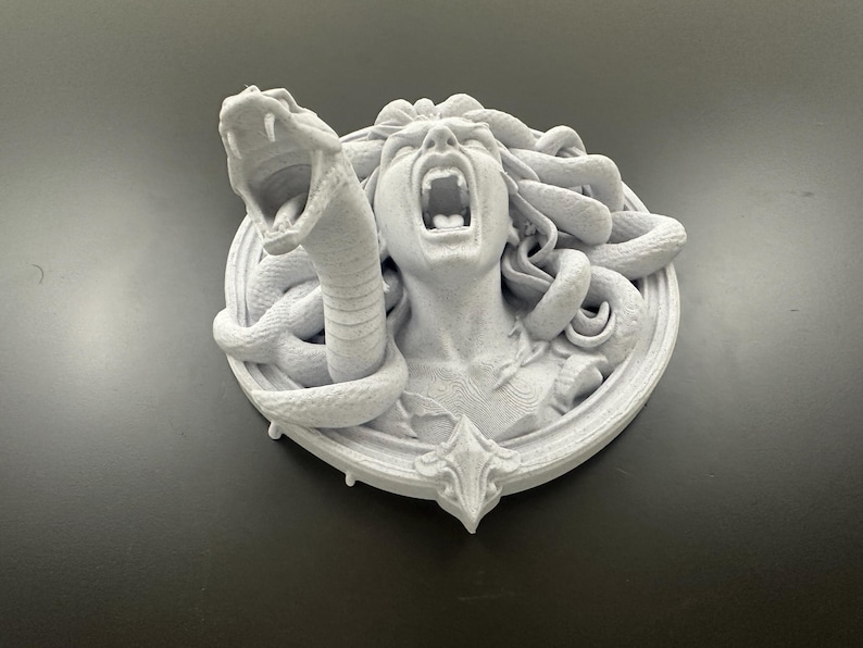 May include: A white sculpture of Medusa with snakes for hair, screaming with her mouth open. A snake is coiled around her head, with its mouth open. The sculpture is mounted on a circular base with a decorative bottom.